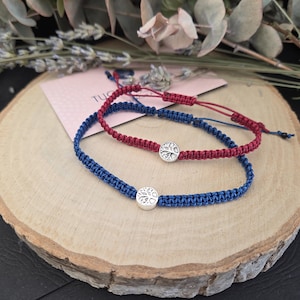 May include: Two macrame bracelets, one blue and one red, each featuring a silver tree of life charm. The bracelets are displayed on a wooden slice, with greenery in the background. The bracelets are adjustable.