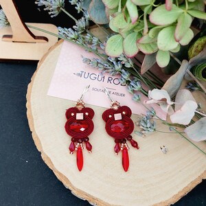 May include: A pair of red soutache earrings with a bear-like design. Each earring features a large, faceted red crystal and dangling red teardrop-shaped beads. The earrings are displayed on a wooden slice with floral accents.