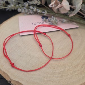 Può includere: Due braccialetti di corda rosso brillante sono esposti su una superficie di legno, con un piccolo biglietto rosa e vegetazione sullo sfondo. I braccialetti sono annodati e sembrano delicati. La carta ha il testo "TUGUI R".