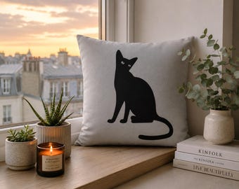 Puede incluir: Un cojín cuadrado de color gris claro que presenta una silueta negra sólida de un gato sentado. El gato está representado de perfil con la cabeza girada hacia el espectador. El cojín tiene una textura de tela lisa y mate con bordes rectos.