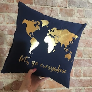 Può includere: Un cuscino blu navy con una mappa del mondo in foglia d'oro e il testo "lets go everywhere" in foglia d'oro.