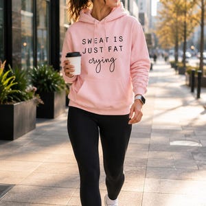 Puede incluir: Una sudadera con capucha de color rosa claro que presenta un bolsillo tipo canguro frontal y el texto negro "SWEAT IS JUST FAT crying" impreso en el pecho. La sudadera se combina con leggings negros lisos, zapatillas deportivas blancas, calcetines blancos y auriculares.