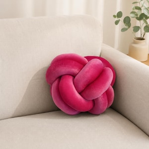 Könnte beinhalten: Ein leuchtend pinkfarbenes Knotenkissen liegt auf einem neutralen Sofa. Das plüschige, geknotete Design schafft einen einzigartigen dekorativen Akzent. Die satte Farbe des Kissens sorgt für einen Hauch von Helligkeit in der Raumdekoration. Es ist ein weiches, dekoratives Element.