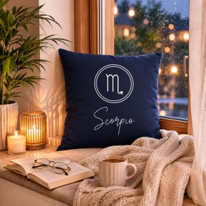 Peut inclure: Un coussin carré bleu marine orné d'un symbole du zodiaque Scorpion blanc dans un cercle et du mot Scorpio écrit en cursive. Le coussin est posé près d'une fenêtre avec un livre, des lunettes, une bougie et une couverture en tricot beige.