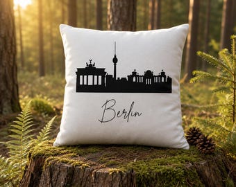 May include: A square white throw pillow featuring a black silhouette of the Berlin skyline, including the Brandenburg Gate and the TV Tower. The word Berlin is printed in a black cursive script font centered below the city skyline graphic.