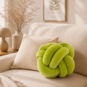 Könnte beinhalten: Ein limettengrünes, knotenförmiges Zierkissen liegt auf einem beigen Sofa. Das Kissen besteht aus einem weichen, plüschigen Material. Ein quadratisches beiges Kissen und eine beige Decke befinden sich ebenfalls auf dem Sofa. Ein gerahmter Druck und eine dekorative Vase sind im Hintergrund.