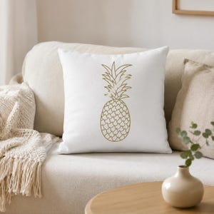 Könnte beinhalten: Ein quadratisches Dekokissen mit weißem Stoffbezug, das eine zentrierte, goldfarbene Strichzeichnung einer Ananas zeigt. Das Design besteht aus einem detaillierten Ananaskörper mit Rautenmuster und einer blättrigen Krone an der Oberseite.