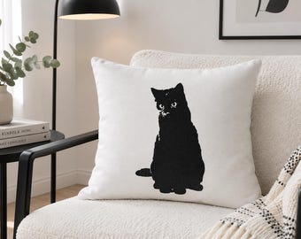 Puede incluir: Un cojín cuadrado de color blanco que presenta una ilustración central en silueta negra sólida de un gato sentado. El diseño del gato está realizado en un estilo gráfico audaz con textura visible, contrastando fuertemente con la tela blanca.