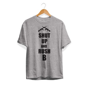 May include: A gray t-shirt with a black graphic of two crossed rifles and the text "SHUT UP AND RUSH B".