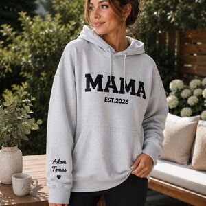May include: A light gray hoodie with the word "MAMA" in bold black letters and "EST. 2026" underneath. The left sleeve has the text "Adam Tomas" and a heart symbol. The hoodie has a front pocket and a drawstring hood.