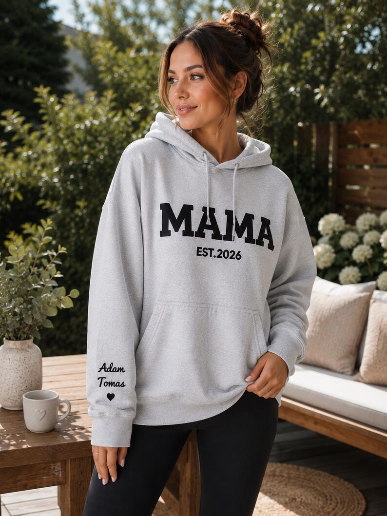 May include: A light gray hoodie with the word "MAMA" in bold black letters and "EST. 2026" underneath. The left sleeve has the text "Adam Tomas" and a heart symbol. The hoodie has a front pocket and a drawstring hood.