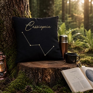 May include: A square black throw pillow featuring the gold-coloured text Cassiopeia and a matching gold constellation graphic. The pillow is positioned on a tree stump alongside a copper-coloured lantern, a metal thermos, a black mug, and an open book.