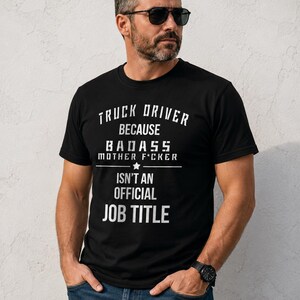 Op de afbeelding: Een zwart t-shirt met korte mouwen en witte tekst die luidt: TRUCK DRIVER BECAUSE BADASS MOTHER F*CKER ISN'T AN OFFICIAL JOB TITLE. Het ontwerp bevat een horizontale lijn met een ster in het midden, geplaatst tussen de tekstregels.