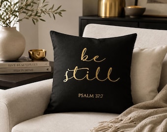 以下が含まれることがあります： 金色の文字で be still と PSALM 37:7 が筆記体でプリントされた、正方形の黒いクッション。明るい色の布張りチェアの上に置かれており、すっきりとシンプルなデザインが特徴です。