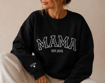May include: Black sweatshirt with the word "MAMA" in white block letters, with "EST. 2013" below. The left sleeve has the name "ZUZIA" and a heart. The sweatshirt is worn with cream-colored sweatpants.