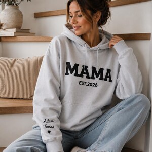 May include: A light gray hoodie with the word "MAMA" in bold black letters. Below the word "MAMA" is the text "EST.2026". The left sleeve has the text "Adam Tomas" and a heart symbol. The person is wearing light blue jeans and white sneakers.