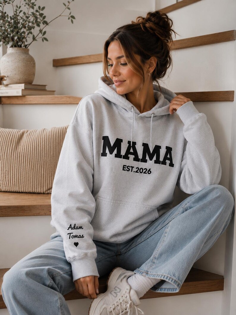 May include: A light gray hoodie with the word "MAMA" in bold black letters. Below the word "MAMA" is the text "EST.2026". The left sleeve has the text "Adam Tomas" and a heart symbol. The person is wearing light blue jeans and white sneakers.