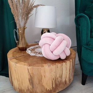 K&ouml;nnte beinhalten: Ein rosa geknotetes Kissen liegt auf einem Holz-Couchtisch mit einer wei&szlig;en Tischdecke. Eine Lampe mit wei&szlig;em Schirm und eine Vase mit getrocknetem Pampasgras stehen auf dem Tisch. Ein gr&uuml;ner Samt-Sessel befindet sich im Hintergrund.