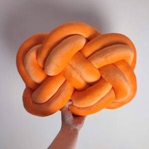 Op de afbeelding: Een groot, oranje, geknoopt kussen. Het kussen is gemaakt van zacht, pluche stof en heeft de vorm van een grote knoop.
