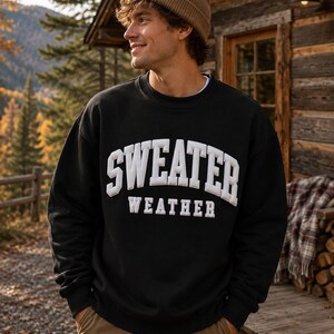 Op de afbeelding: Een zwart sweatshirt met lange mouwen en ronde hals, voorzien van de witte tekst SWEATER WEATHER in een dik, verhoogd lettertype in collegestijl op de borst, gecombineerd met een beige gebreide muts en een bruine broek voor een casual uitstraling.