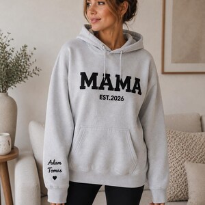 May include: A light gray hoodie with the word "MAMA" in bold black letters and "EST.2026" underneath. The left sleeve has the text "Adam Tomas" and a heart symbol. The hoodie has a front pocket and a drawstring hood.