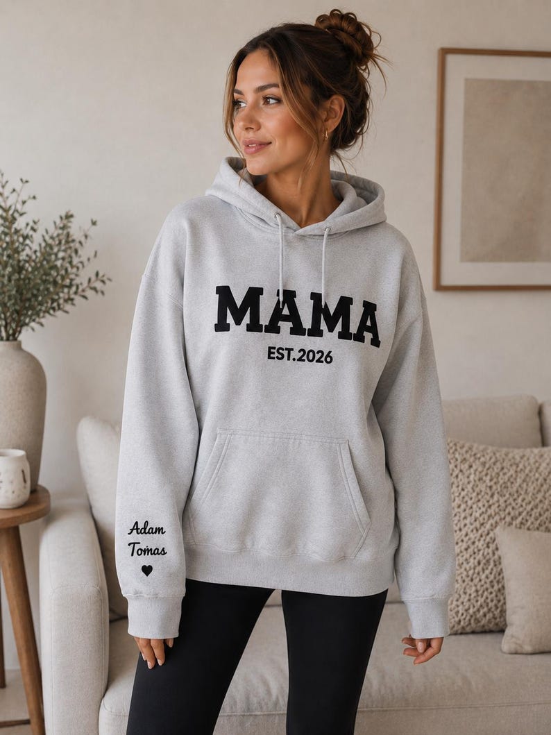May include: A light gray hoodie with the word "MAMA" in bold black letters and "EST.2026" underneath. The left sleeve has the text "Adam Tomas" and a heart symbol. The hoodie has a front pocket and a drawstring hood.