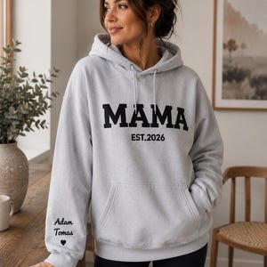 May include: A light gray hoodie with the word "MAMA" in bold black letters, and "EST.2026" below. The left sleeve has the names "Adam Tomas" and a heart symbol. The hoodie has a front pocket and a drawstring hood.