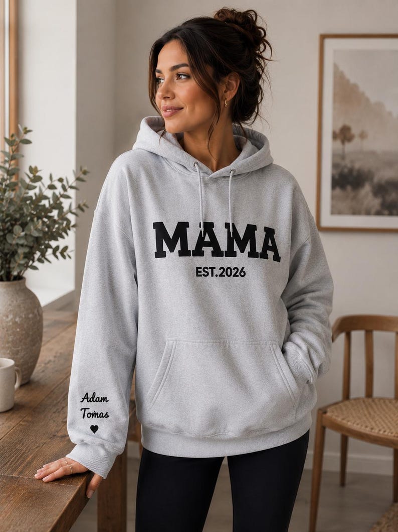 May include: A light gray hoodie with the word "MAMA" in bold black letters, and "EST.2026" below. The left sleeve has the names "Adam Tomas" and a heart symbol. The hoodie has a front pocket and a drawstring hood.