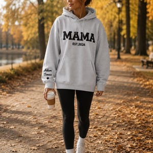 May include: A light gray hoodie with the word "MAMA" in bold black letters and "EST.2026" underneath. The left sleeve has the text "Adam Tomas" and a heart. The person is wearing black leggings and white sneakers.