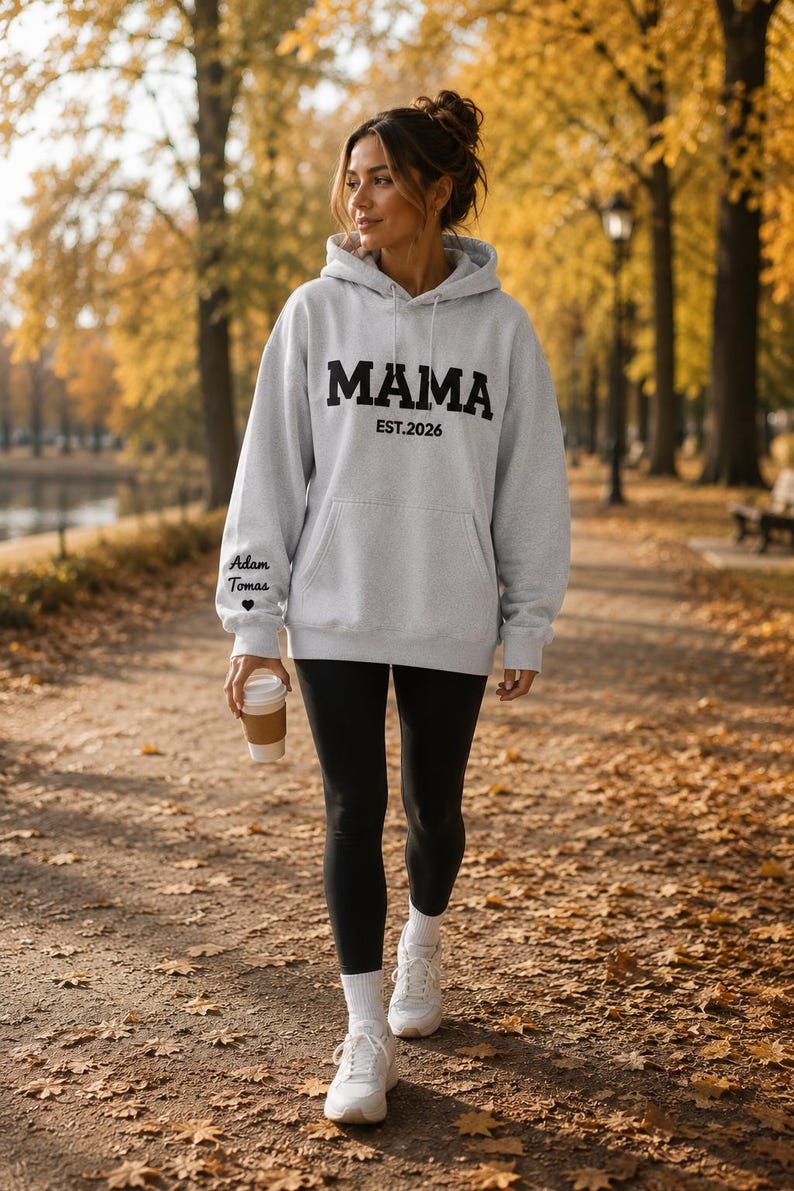 May include: A light gray hoodie with the word "MAMA" in bold black letters and "EST.2026" underneath. The left sleeve has the text "Adam Tomas" and a heart. The person is wearing black leggings and white sneakers.