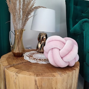 K&ouml;nnte beinhalten: Ein Holz-Couchtisch mit einem Knotenkissen aus rosa Samt darauf. Eine wei&szlig;e Lampe mit goldener Basis und eine Vase mit getrocknetem Pampasgras stehen ebenfalls auf dem Tisch.