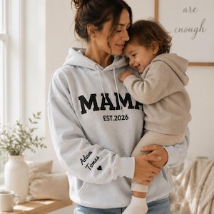 May include: A light gray hoodie with the word "MAMA" in bold black letters and "EST. 2026" printed below. The left sleeve has the names "Adam Tomas" and a heart symbol. The hoodie is worn by a person holding a child.