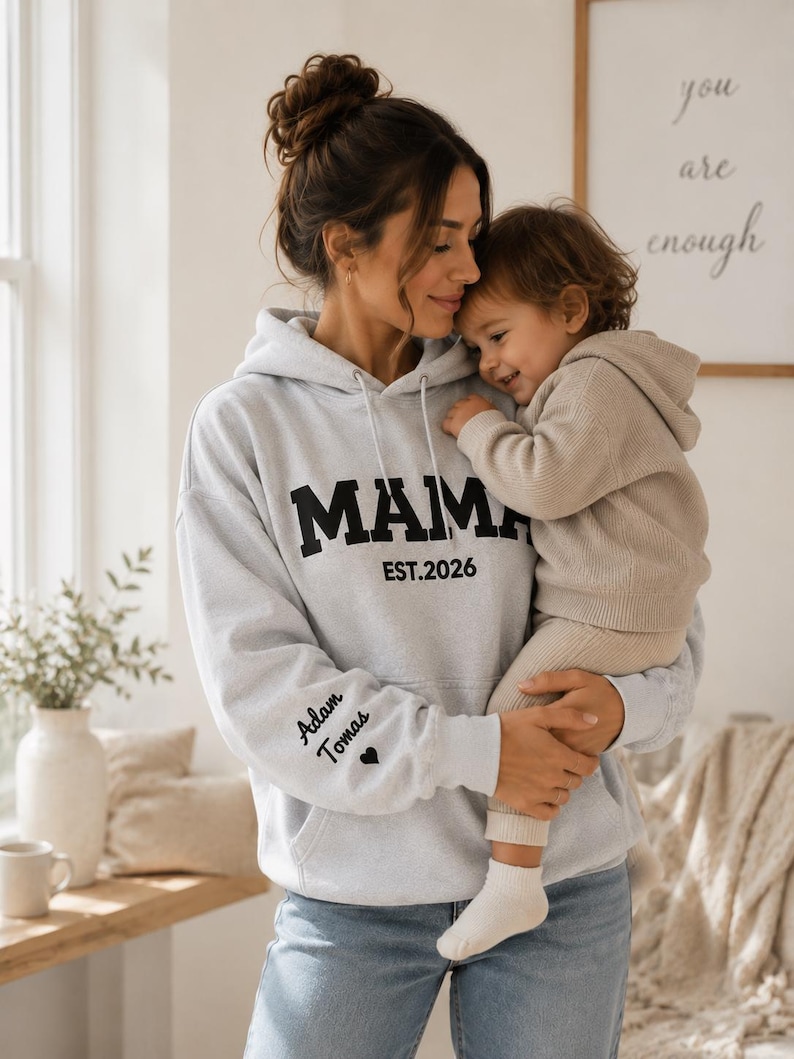 May include: A light gray hoodie with the word "MAMA" in bold black letters and "EST. 2026" printed below. The left sleeve has the names "Adam Tomas" and a heart symbol. The hoodie is worn by a person holding a child.