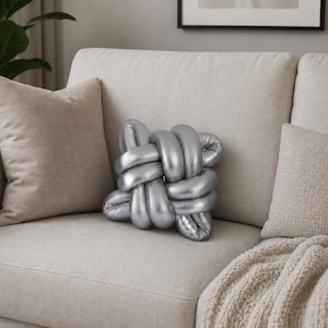 May include: A silver decorative knot pillow rests on a beige sofa, flanked by two neutral-toned throw pillows. The knot pillow has a metallic sheen and a unique, intertwined design, adding a modern touch to the living room setting.