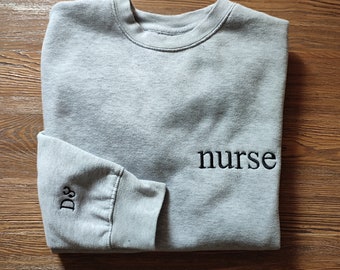 May include: A gray sweatshirt with the word "nurse" embroidered on the front in black. The word "DS" is embroidered on the left sleeve in black.