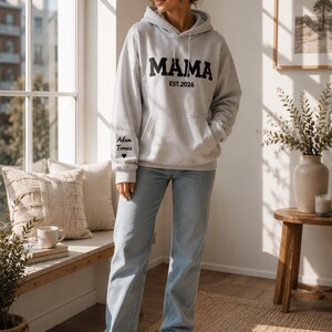 May include: A light gray hoodie with the word "MAMA" in bold black letters and "EST.2026" printed below. The left sleeve has the names "Adam Tomas" and a heart symbol. The person is wearing light blue jeans and white sneakers.