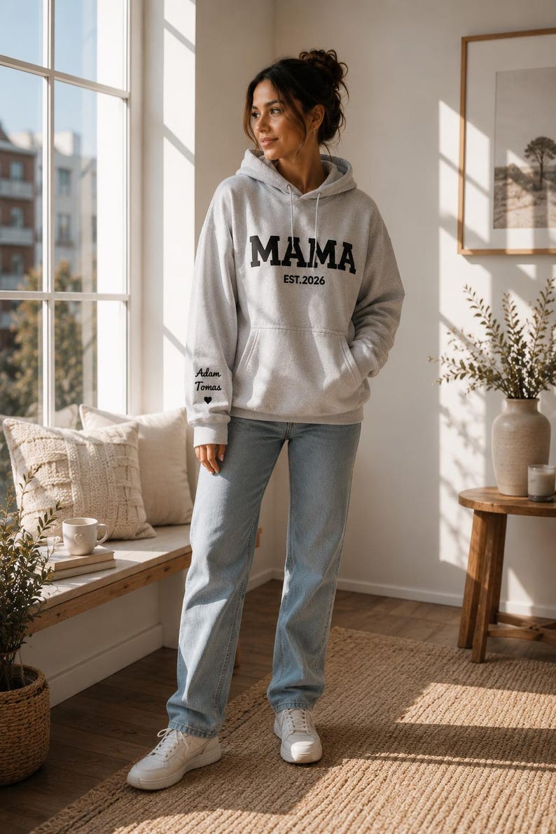 May include: A light gray hoodie with the word "MAMA" in bold black letters and "EST.2026" printed below. The left sleeve has the names "Adam Tomas" and a heart symbol. The person is wearing light blue jeans and white sneakers.