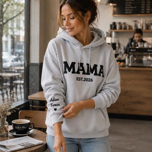 May include: A light gray hoodie with the word "MAMA" in bold black letters and "EST.2026" printed below. The left sleeve has the text "Adam Tomas" and a heart symbol. The hoodie is worn with blue jeans.