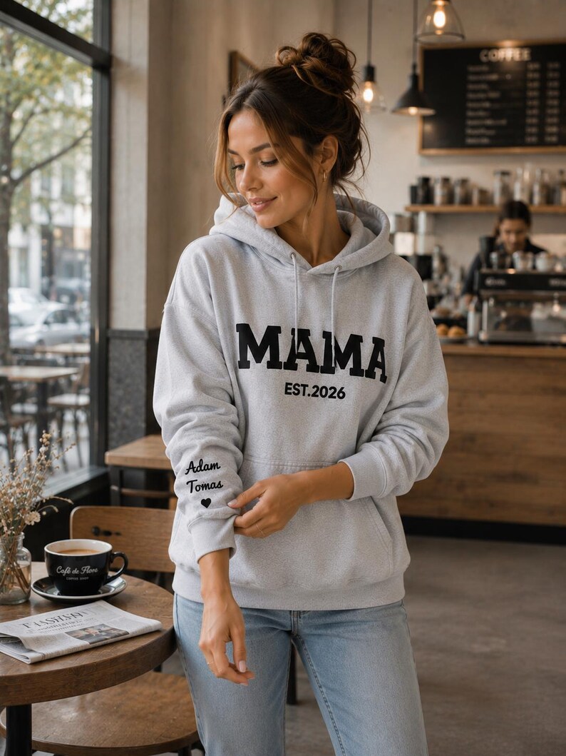 May include: A light gray hoodie with the word "MAMA" in bold black letters and "EST.2026" printed below. The left sleeve has the text "Adam Tomas" and a heart symbol. The hoodie is worn with blue jeans.