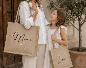 May include: Two rectangular jute tote bags with sturdy handles. The larger bag features the black script text Mama and a small gold heart. The smaller bag features the black script text Lena and a small gold heart. Both bags have a natural tan color.