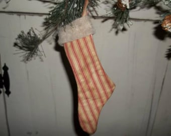 May include: A Christmas stocking with a cream-colored background and red vertical stripes. The top of the stocking has a fluffy, cream-colored trim. The stocking is hanging from a green branch with pine needles and small pine cones.