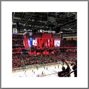Könnte beinhalten: Ein großes rot-weißes Scoreboard in einer Eishockeyarena zeigt die Worte "Devils Win" in roten Buchstaben. Das Scoreboard hängt von der Decke über einer Menge von Menschen, die rote und schwarze Kleidung tragen. Die Eisbahn ist im Vordergrund sichtbar.