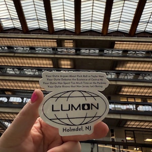 May include: A white sticker with the word "LUMON" in bold black letters, encircled by a globe design. Below, the text "Holmdel, NJ" is printed. Above, a speech bubble contains text about New Jersey. The background features a building with a glass ceiling.