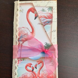May include: A decorative card featuring flamingo illustrations. The card is adorned with a pink ribbon and lace trim. The flamingo illustrations are in shades of pink and red, set against a backdrop of tropical scenery.