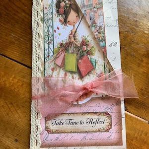 May include: A handmade greeting card with a floral design and a ribbon. The card features an illustration of a woman holding shopping bags. The text "Take Time to Reflect" is displayed on the card. The card is trimmed with lace.