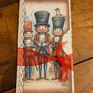 May include: A decorative card featuring three illustrated nutcracker soldiers in red, blue, and gold uniforms. The card is tied with a red ribbon and has a lace trim on the left side. The background shows a winter scene.