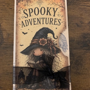 May include: A rectangular card titled Spooky Adventures featuring a drawing of a character in a large black hat reading a book, with a lantern nearby, bats, and a spiderweb. The card is wrapped in a black sheer ribbon with a bow at the bottom.