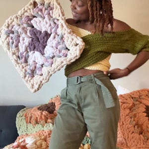 Può includere: Una grande coperta quadrata a maglia grossa nei toni del crema, lavanda e grigio viene tenuta in alto. La coperta ha un aspetto strutturato e fatto a mano. Altre coperte in colori simili sono sullo sfondo. Il tema generale è l'arredamento della casa.