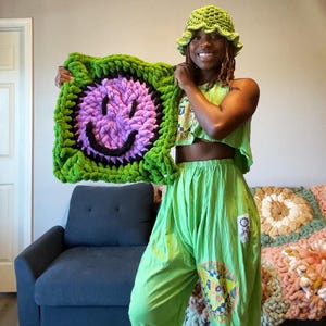 May include: A person wearing a green crochet hat and green pants holds a green and purple crocheted pillow with a black smiley face.