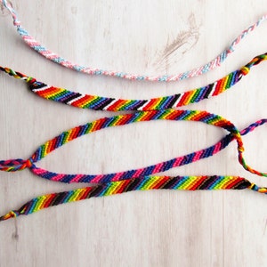 May include: Four handmade friendship bracelets made of woven embroidery floss. One bracelet features a light blue, pink, and white twisted pattern. Three bracelets display diagonal stripes in vibrant rainbow colors, including red, orange, yellow, green, blue, and purple.